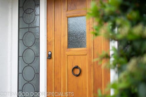 1930s style front door with small window. Natural finish wooden door. Black iron door furniture giving a rustic or Tudor appearance. Obscured glass to small window in door, frame with leaded Art Deco geometric design.