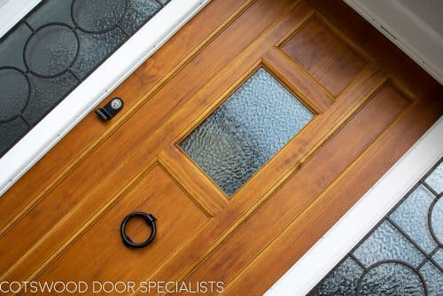 1930s style front door with small window. Natural finish wooden door. Black iron door furniture giving a rustic or Tudor appearance. Obscured glass to small window in door, frame with leaded Art Deco geometric design.
