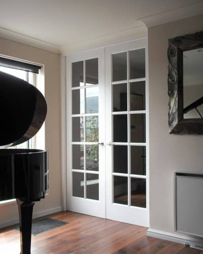 Georgian glazed internal doors with door frame. Extra tall doors painted white with clear glass. Double doors with narrow glazing bar. Light shining on doors in room with grand piano