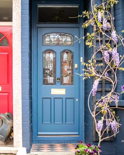 Stained Glass Victorian door fitted into classic London terraced house. Three arched top stained glass panels and three wooden panels. Special gold leaf number above door. Door, frame and house painted blue in farrow and ball colours. Wisteria growing around front door. Polished brass door furniture including letterbox and knob