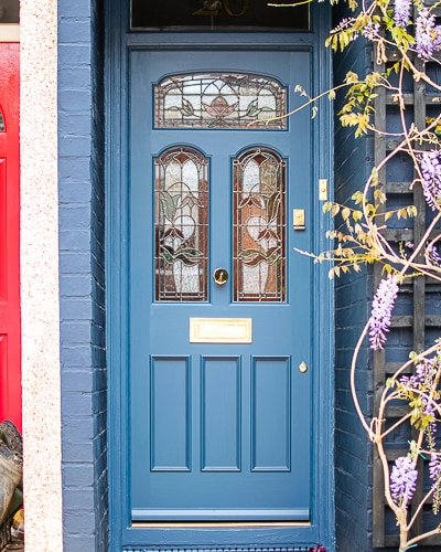 Stained Glass Victorian door fitted into classic London terraced house. Three arched top stained glass panels and three wooden panels. Special gold leaf number above door. Door, frame and house painted blue in farrow and ball colours. Wisteria growing around front door. Polished brass door furniture including letterbox and knob