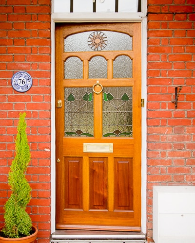 stained timber Edwardian front door. Edwardian design stained glass including a sun pattern. Polished brass door furniture. Door fitted into an existing painted white door frame surrounded by red brickwork