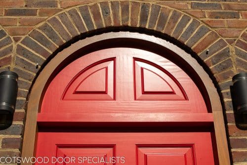 Red Georgian 5 panel front door fitted into an arched door frame. Solid door and frame with no glass painted bright red. Raised solid door panels. Polished brass door furniture. Closeup of arched door frame and panel