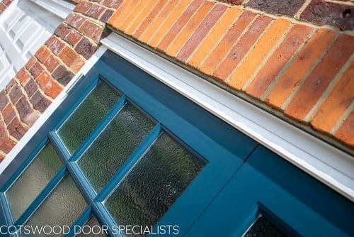 1930s wooden garage doors with matching front door. Fitted into a 1930s house with red brickwork. Doors painted dark blue. Front door has stained glass, garage doors have traditional textured obscure glass. Satin chrome door hardware. Closeup of glazing bar and head drip detail