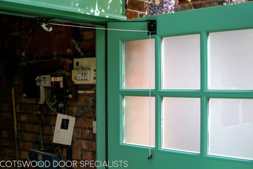 1930s art deco garage doors. Classic 1930's side hinged garage doors with glazing. Glazing is divided into 6 pieces per door with wooden glazing bar. Obscured glass. Painted light green. Tongue and groove style boarding. polished chrome door furniture. Close up of door holder.