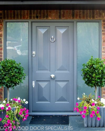 Contemporary Victorian front door painted grey. Door has 4 bold cricket bat panels and is fitted into a sidelight frame. Frame has etched glass. Picture from front of house with plants surrounding door. House is red brick. Wooden door is fitted with stain chrome banham locks and door furniture
