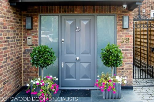 Contemporary Victorian front door painted grey. Door has 4 bold cricket bat panels and is fitted into a sidelight frame. Frame has etched glass. Picture from front of house with plants surrounding door. House is red brick. Wooden door is fitted with stain chrome banham locks and door furniture.