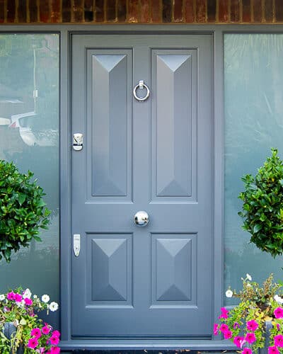 Contemporary Victorian front door painted grey. Door has 4 bold cricket bat panels and is fitted into a sidelight frame. Frame has etched glass. Picture from front of house with plants surrounding door. House is red brick. Wooden door is fitted with stain chrome banham locks and door furniture