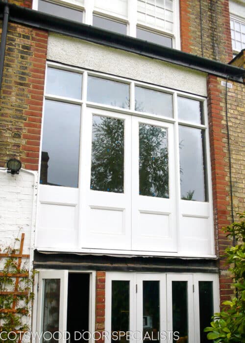 Period Juliet balcony doors fitted into a kitchen. White painted doors half glazed with windows above doors. Clear glass looking out to garden.