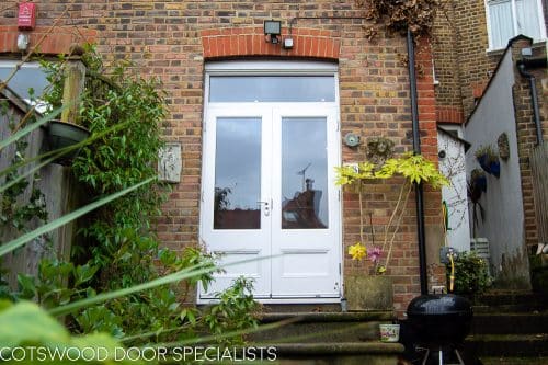 Wooden French doors fitted into a London home. Doors leading out to the garden from kitchen. Flat panels to bottom of doors with traditional panel mouldings. Doors and frame double glazed and painted white