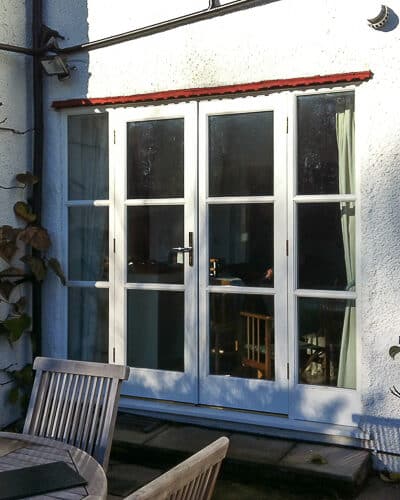 1930's Art Deco French door. Wooden doors and frame painted white with horizontal glazing bars. Clear double glazed glass. Doors fitted into white rendered house with 1930s creasing tile detail. Doors open onto patio with seating area