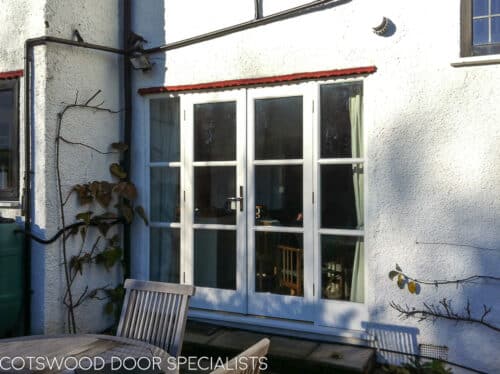 1930's Art Deco French door. Wooden doors and frame painted white with horizontal glazing bars. Clear double glazed glass. Doors fitted into white rendered house with 1930s creasing tile detail. Doors open onto patio with seating area
