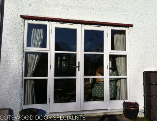 1930s French door with opening windows. Wooden French door fully painted white with clear double glazed glass. Doors and side windows leading out onto garden. Brass door handles. Doors viewed from outside with clean white render and classic 1930s creasing tile detail above doors