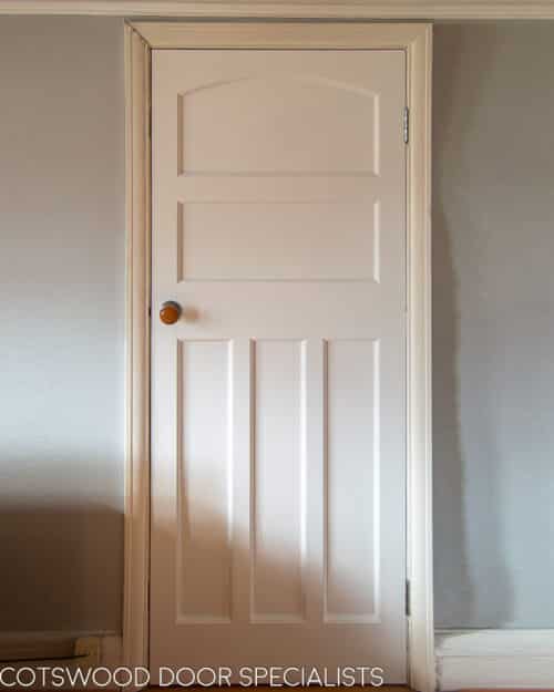 1930s Internal door. The door has multiple recessed panels with three long vertical panels and a shaped panel at the top of the door. Door is painted white and installed with a wooden door knob