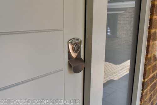 Contemporary external door. Wide wooden door with horizontal boards painted in a green grey colour. Etched glass to sidelights. Satin chrome door furniture. Closeup of lock