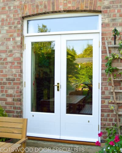 French patio doors. Doors and frame fitted into a london redbrick house, leading out onto patio. Fully painted in white paint. Doors have a low butt and beaded panel. Clear double glazed glass to doors and fanlight. Spring flowers on patio