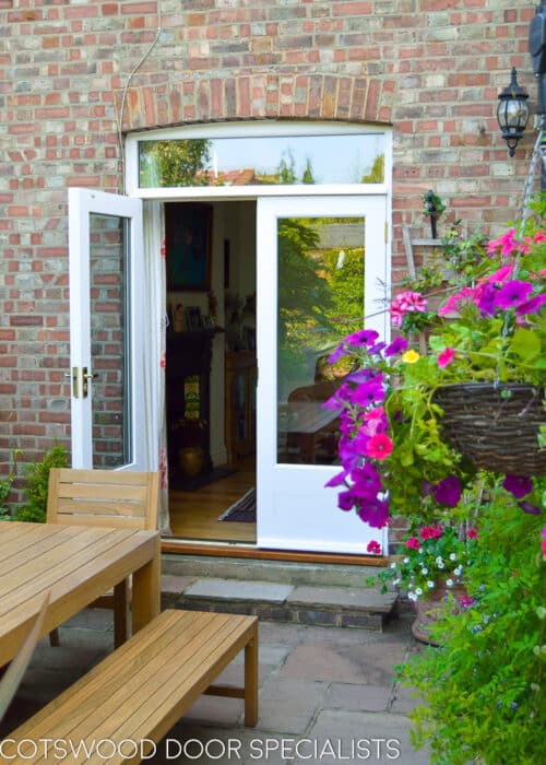 French patio doors. Doors and frame fitted into a london redbrick house, leading out onto patio. Fully painted in white paint. Doors have a low butt and beaded panel. Clear double glazed glass to doors and fanlight. Spring flowers on patio. One door open