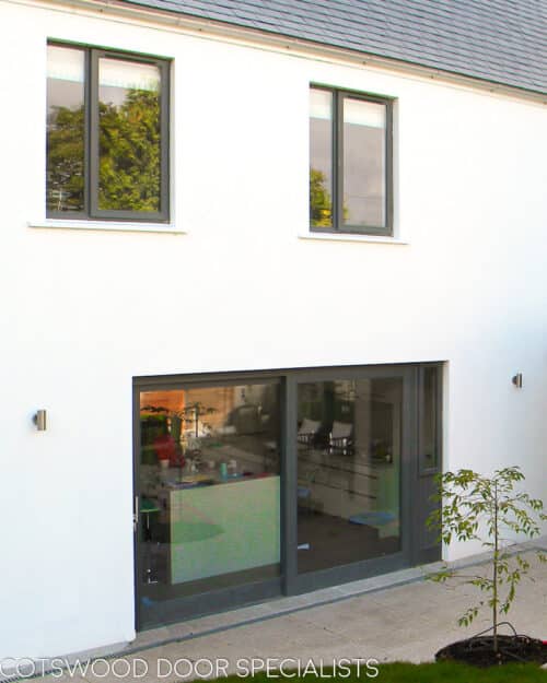 Sliding external wooden door. Grey painted wooden doors with clear double glazed units. Fitted into a contemporary home with white rendered walls. Doors leading out onto patio area