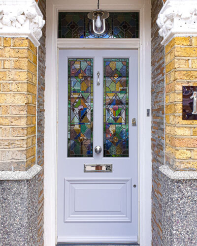 Victorian external door. Door and frame has ornate geometric stained glass. Door has 2 glass panels and one raised wooden panel. Painted finish Victorian door fitted in a storm porch. Polished chrome door furniture