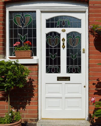 White Edwardian Door. Door has 3 shaped stained glass panes and is painted white. Red Edwardian London home. Front entrance has a glazed wing window also with stained glass. White paint with brass hardware