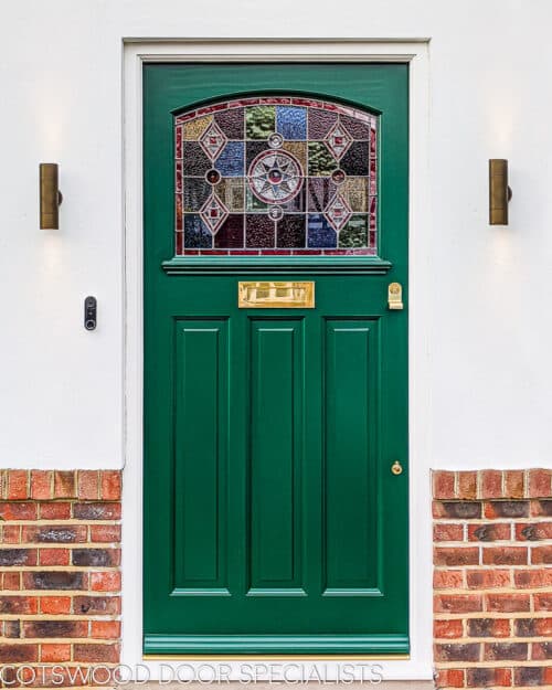 Green 1930s front door fitted with geometric stained glass. Wooden front door fitted into a part brick part rendered home. Door has brass door furniture and coloured glass