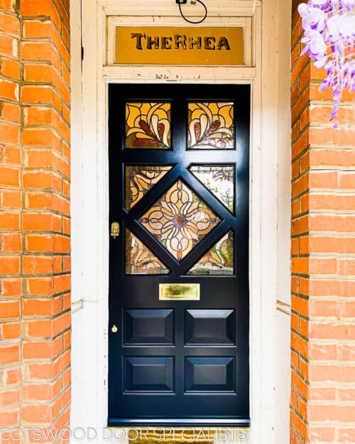 Period Edwardian door. Wooden edwardian front door with diamond shaped stained glass. Period stained glass design with colours and textures of glass. Door painted in a dark blue sympathetic to the surrounding red brick and wisteria plant. Polished brass door furniture