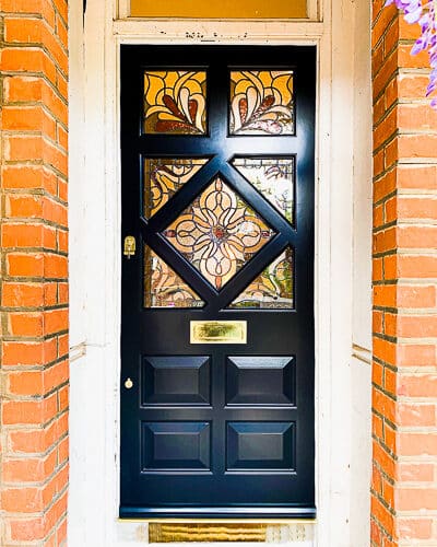 Period Edwardian door. Wooden edwardian front door with diamond shaped stained glass. Period stained glass design with colours and textures of glass. Door painted in a dark blue sympathetic to the surrounding red brick and wisteria plant. Polished brass door furniture
