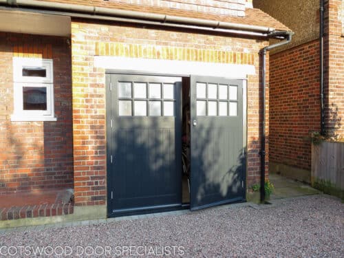 Side hung garage doors. Large grey painted 1930s style garage door with glazed top. Multiple panes of obscured glass divided with wooden glazing bar. Boarded bottom section to doors. Doors fitted with satin chrome door furniture