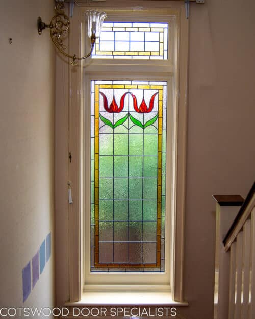 Stained glass casement window fitted onto landing in a large Edwardian home. Stained glass to window has flower motif in red colours. Window is painted white. Internal shot from hallway showing stained glass to window