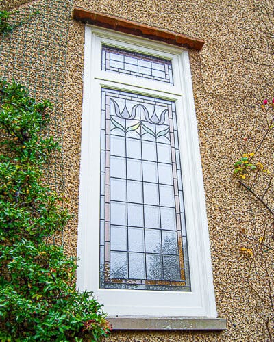 Stained glass casement window fitted onto landing in a large Edwardian home. Stained glass to window has flower motif in red colours. Window is painted white