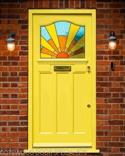 Sunrise 1930s front door. Door and frame painted yellow with an Art deco stained glass sunrise. Polished brass door furniture. Yellow and amber textured glass to sunrise with blue sky. Decorative shelf and apron to door with dental block