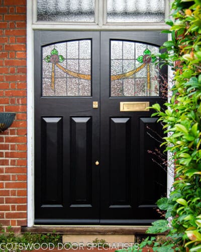 Black 1930's front doors, double doors featuring stained glass fitted into a semi detached home. White surrounding door frame and red brickwork. Stained glass has a rose design with green petals. Polished brass letterbox