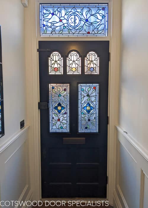 Classic blue Victorian door with shaped glazing. Stained glass design with leaves and beautiful colours. Decoraitve rondels as centre features in different colours. Number to transom panel. Door is a very dark blue and is fitted with bronze door furniture including banham locks. Interior hallway shot showing off stained glass