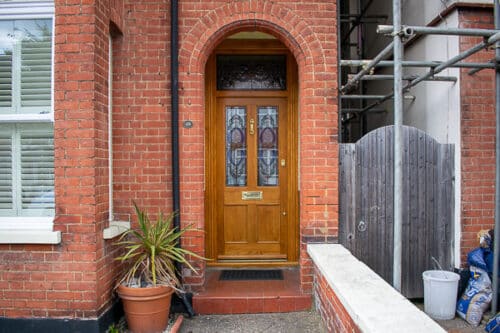 Natural Victorian Door. Natural finish Accoya wood front door and door frame fitted into a red brick London home. Translucent finish shows off the grain of Accoya wood. Traditional stained glass to door and frame above. Door has 4 panels, of which the top 2 are glazed. Polished brass hardware including a doctor knocker