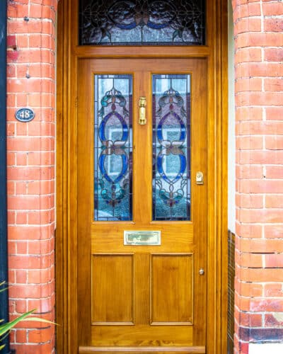 Natural Victorian Door. Natural finish Accoya wood front door and door frame fitted into a red brick London home. Translucent finish shows off the grain of Accoya wood. Traditional stained glass to door and frame above. Door has 4 panels, of which the top 2 are glazed. Polished brass hardware including a doctor knocker