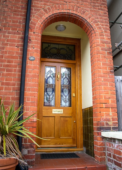Natural Victorian Door. Natural finish Accoya wood front door and door frame fitted into a red brick London home. Translucent finish shows off the grain of Accoya wood. Traditional stained glass to door and frame above. Door has 4 panels, of which the top 2 are glazed. Polished brass hardware including a doctor knocker