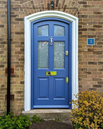 arched Georgian front door painted blue fitted into a brick house. Door and frame are both Accoya wood and painted blue. Banham high security locks. All door hardware polished chrome. Sandblasted glass with Clear inset border, double glazed