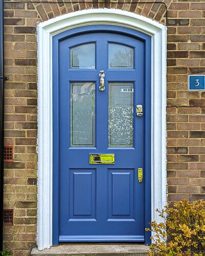 arched Georgian front door painted blue fitted into a brick house. Door and frame are both Accoya wood and painted blue. Banham high security locks. All door hardware polished chrome. Sandblasted glass with Clear inset border, double glazed