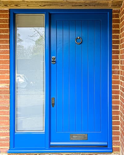Blue contemporary front door with sidelight frame . Door is made in a tradional way with vertical boards. Frame has a full height piece of glass that is sandblasted with a clear inset line border. Door has dark bronze banham locks, letterplate and knocker. Door and frame painted blue