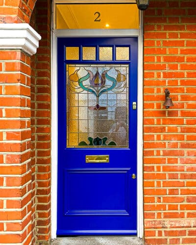 Floral Edwardian front door. Painted blue Edwardian front dooor with elegant floral design stained glass. Polished brass door hardware. Door is fitted into a red brick house. Stained glass has different textures and a reeded border.