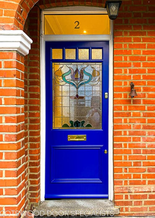 Floral Edwardian front door. Painted blue Edwardian front door with elegant floral design stained glass. Polished brass door hardware. Door is fitted into a red brick house. Stained glass has different textures and a reeded border.