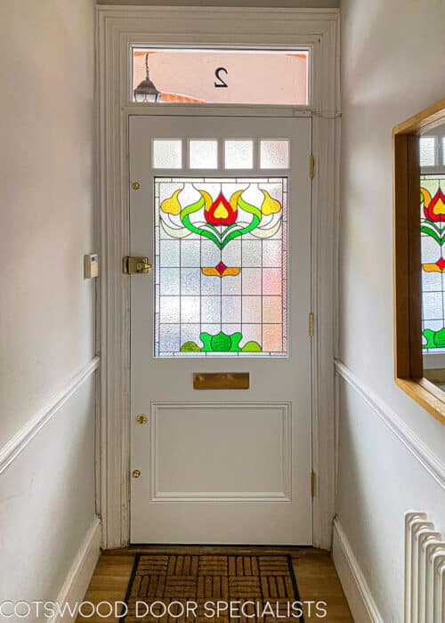Floral Edwardian front door. Painted blue Edwardian front door with elegant floral design stained glass. Polished brass door hardware. Door is fitted into a red brick house. Stained glass has different textures and a reeded border. Internal hallway photo showing off stained glass