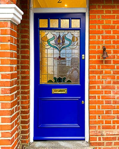 Floral Edwardian front door. Painted blue Edwardian front door with elegant floral design stained glass. Polished brass door hardware. Door is fitted into a red brick house. Stained glass has different textures and a reeded border.