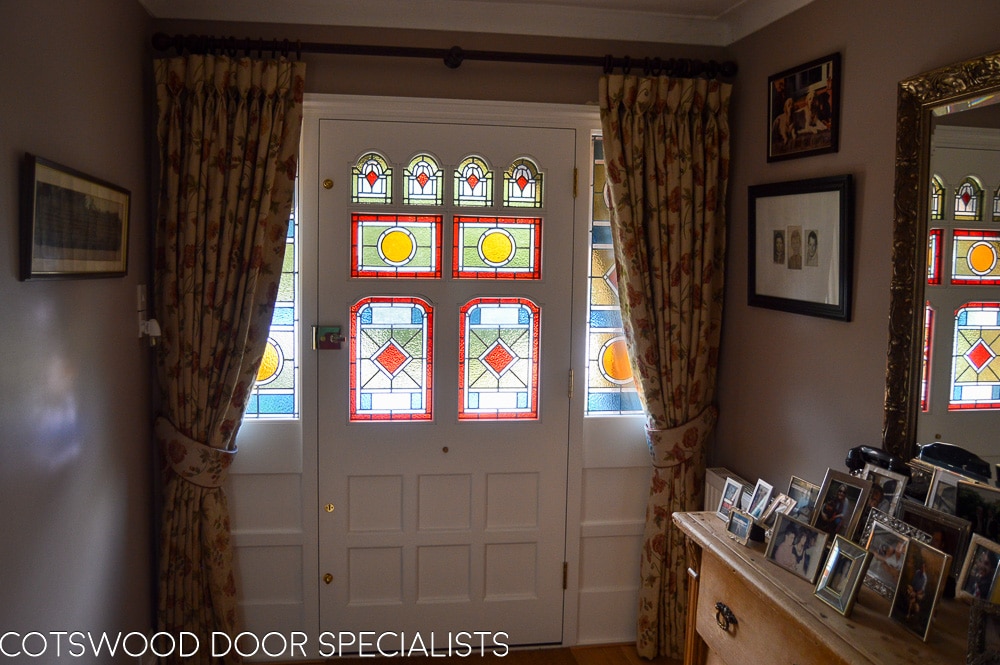 grey painted Victorian front door and double sidelight frame fitted in london. door has vibrant geometric stained glass. laminated stained glass. Draught proofed and insurance rated locks fitted. door furniture is polished brass. bright colours flood light into hallway though glass. Door is of an intricate design with 8 panes of glass, some of which are shaped. Hallway shot showing stained glass