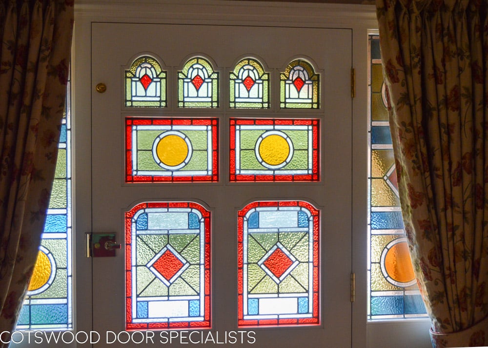 grey painted Victorian front door and double sidelight frame fitted in london. door has vibrant geometric stained glass. laminated stained glass. Draught proofed and insurance rated locks fitted. door furniture is polished brass. bright colours flood light into hallway though glass. Door is of an intricate design with 8 panes of glass, some of which are shaped. Hallway shot showing stained glass